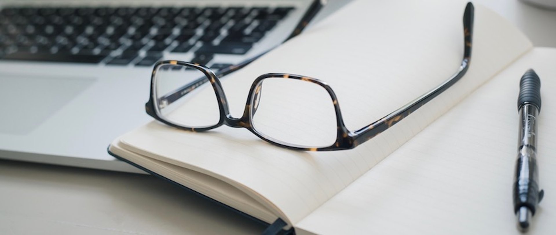 Eyeglasses resting on an open notebook next to a laptop keyboard and pen on a desk