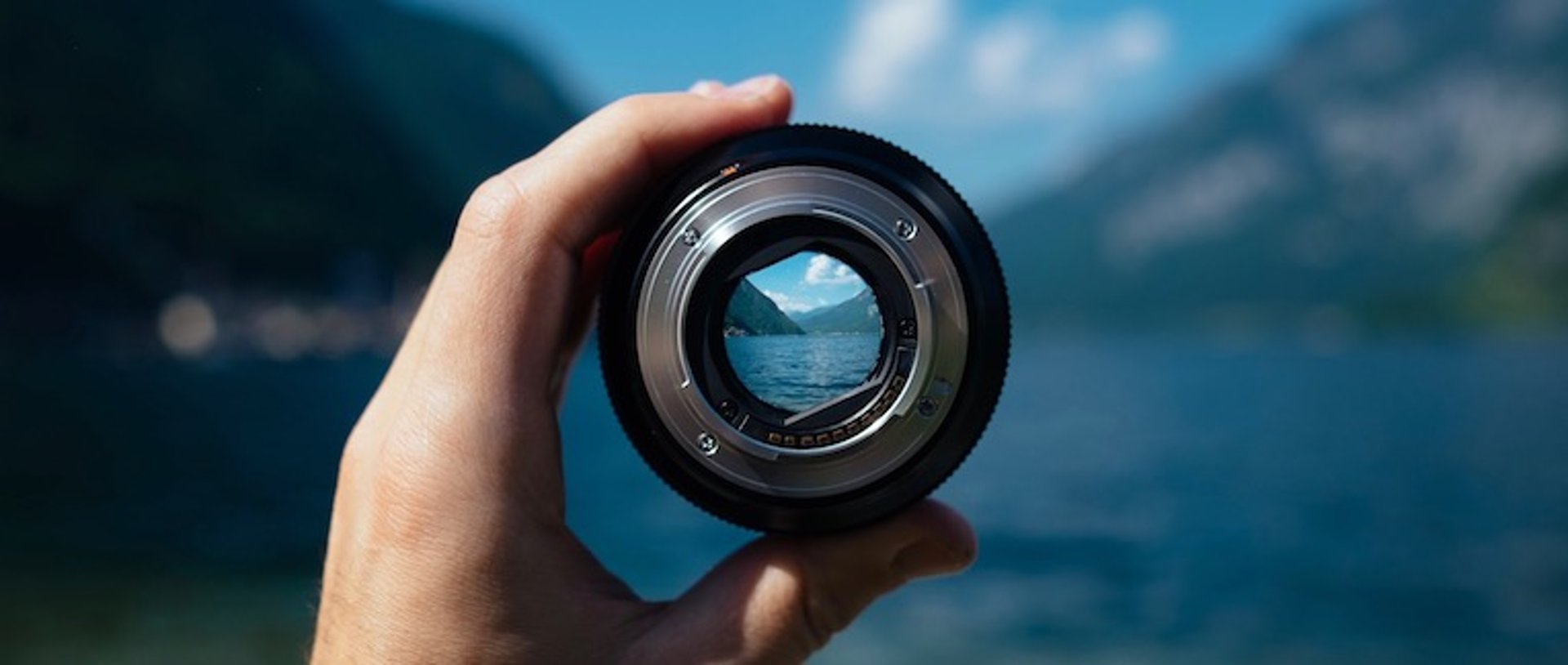Hand holding a camera lens focused on a mountain lake with blue sky and peaks in the background